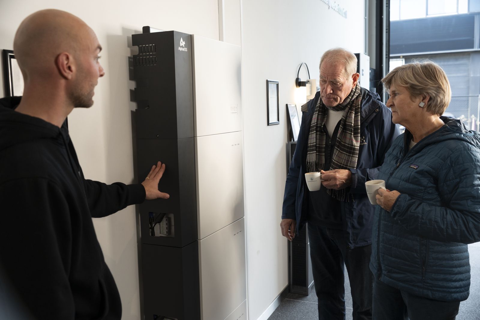 Bezoekers met koffie bekijken een AlphaESS-thuisbatterij terwijl de adviseur uitleg geeft